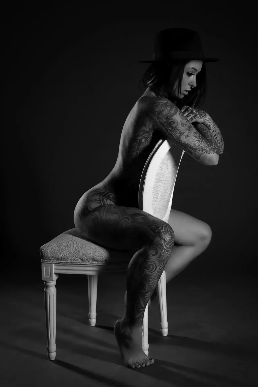 Tätowierte Frau mit Hut auf Stuhl im Profil, monochromes Kunstfoto.