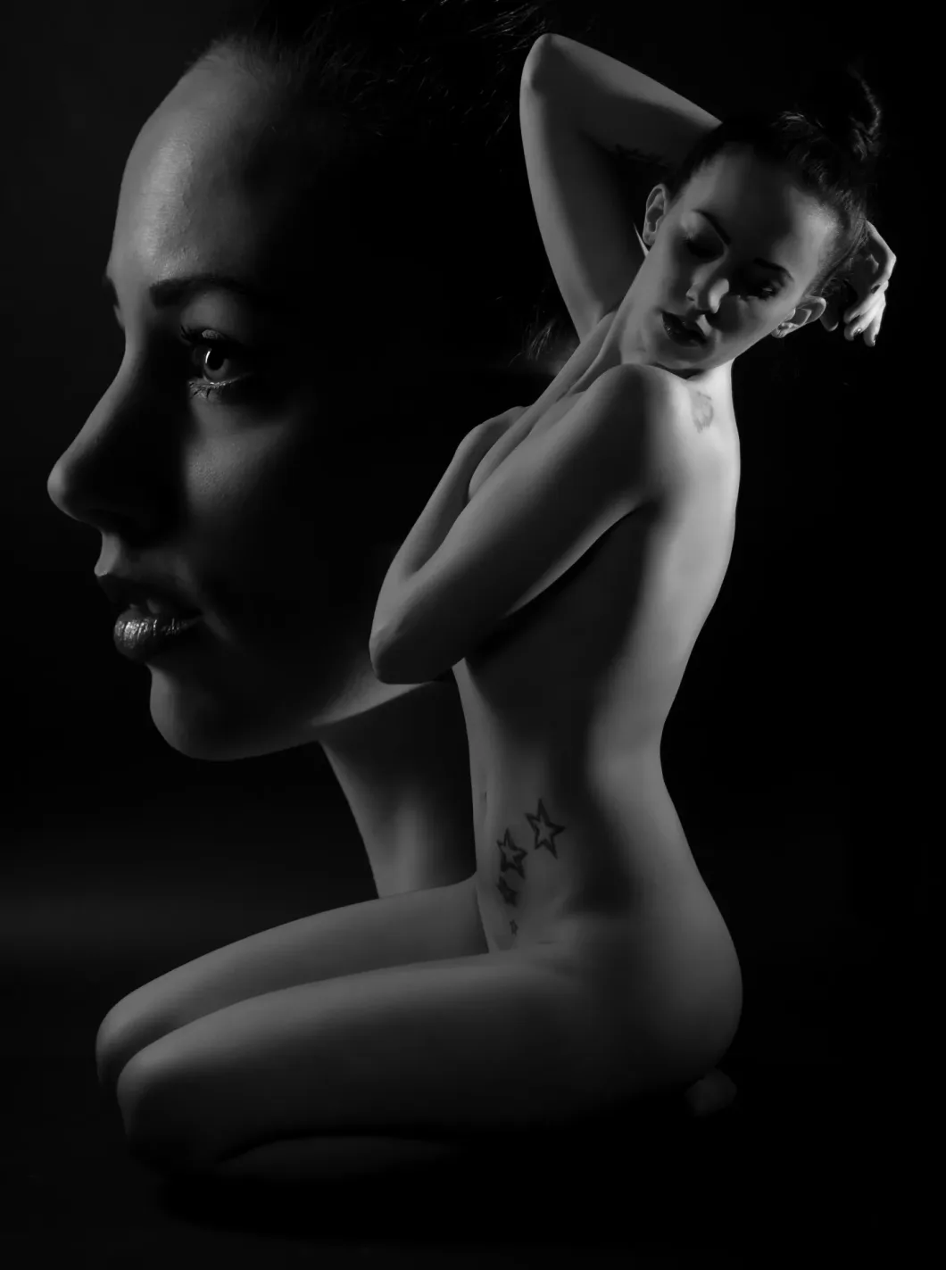 Schwarzweiß-Porträt einer nackten Frau in nachdenklicher Pose mit Tattoos.