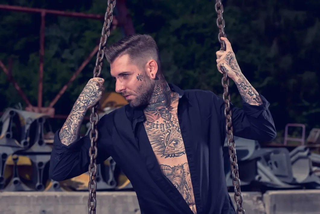 Mann mit Tattoos an Ketten vor industriellem Hintergrund, arrogant posierend.
