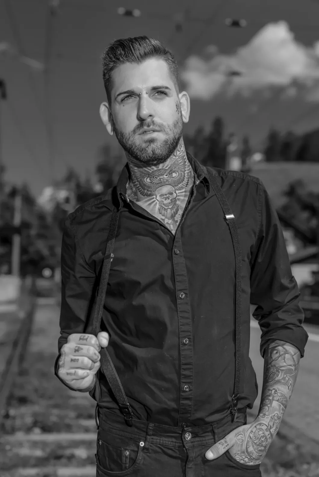Mann mit Tattoos und schwarzen Hemd auf leerer Bahnstrecke, im Hintergrund Bäume und Himmel.