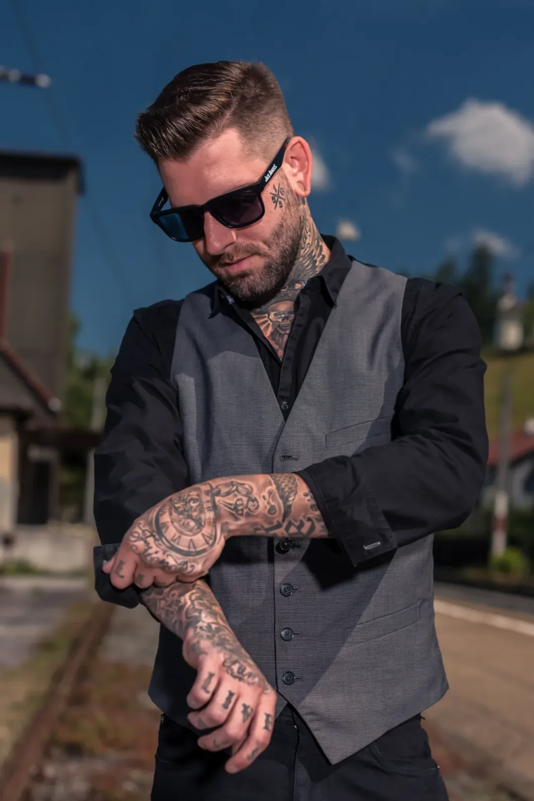Mann mit Sonnenbrille und Tattoos, trägt Weste, steht auf Bahngleisen.