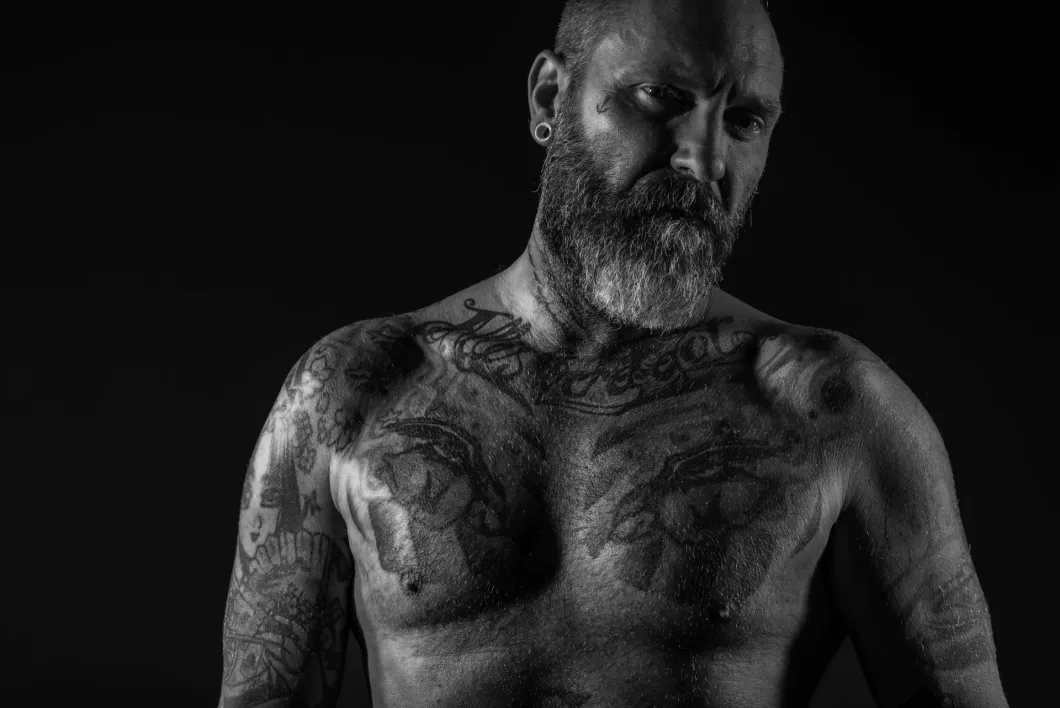 Mann mit Bart und Tattoos auf dunklem Hintergrund, starker Ausdruck, Schwarz-Weiß-Aufnahme.