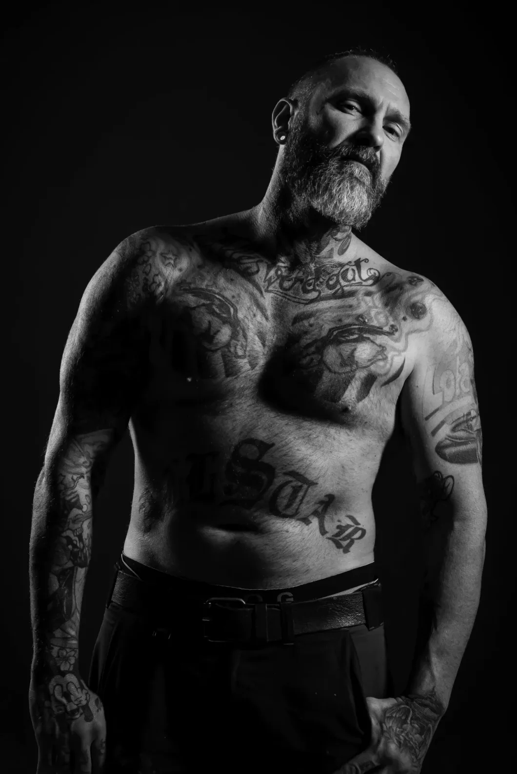 Mann mit Tattoos posiert oberkörperfrei vor dunklem Hintergrund.