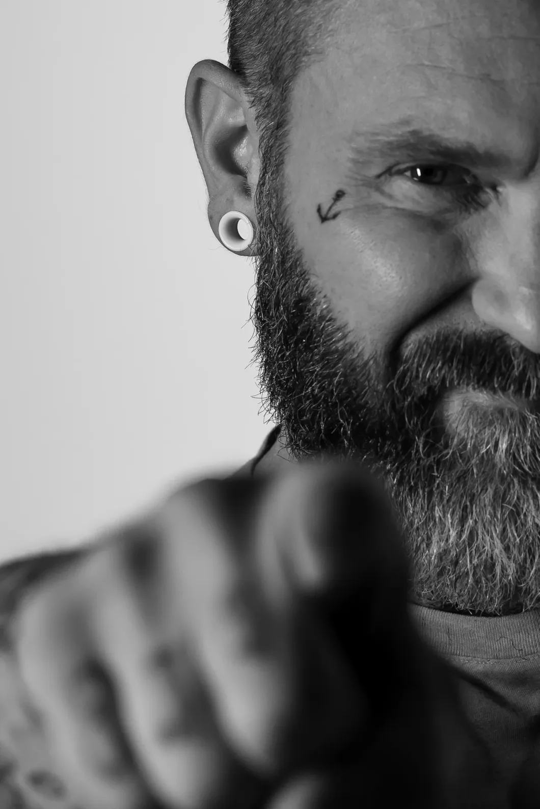 Mann mit Bart zeigt auf Kamera, Nahaufnahme in Schwarz-Weiß, Tattoo im Gesicht sichtbar.