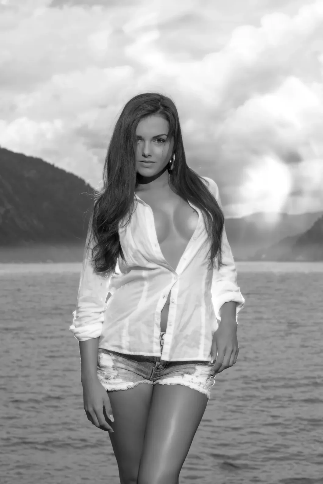 Frau in weißem Hemd und Shorts am See mit Bergen im Hintergrund, schwarzweiß.
