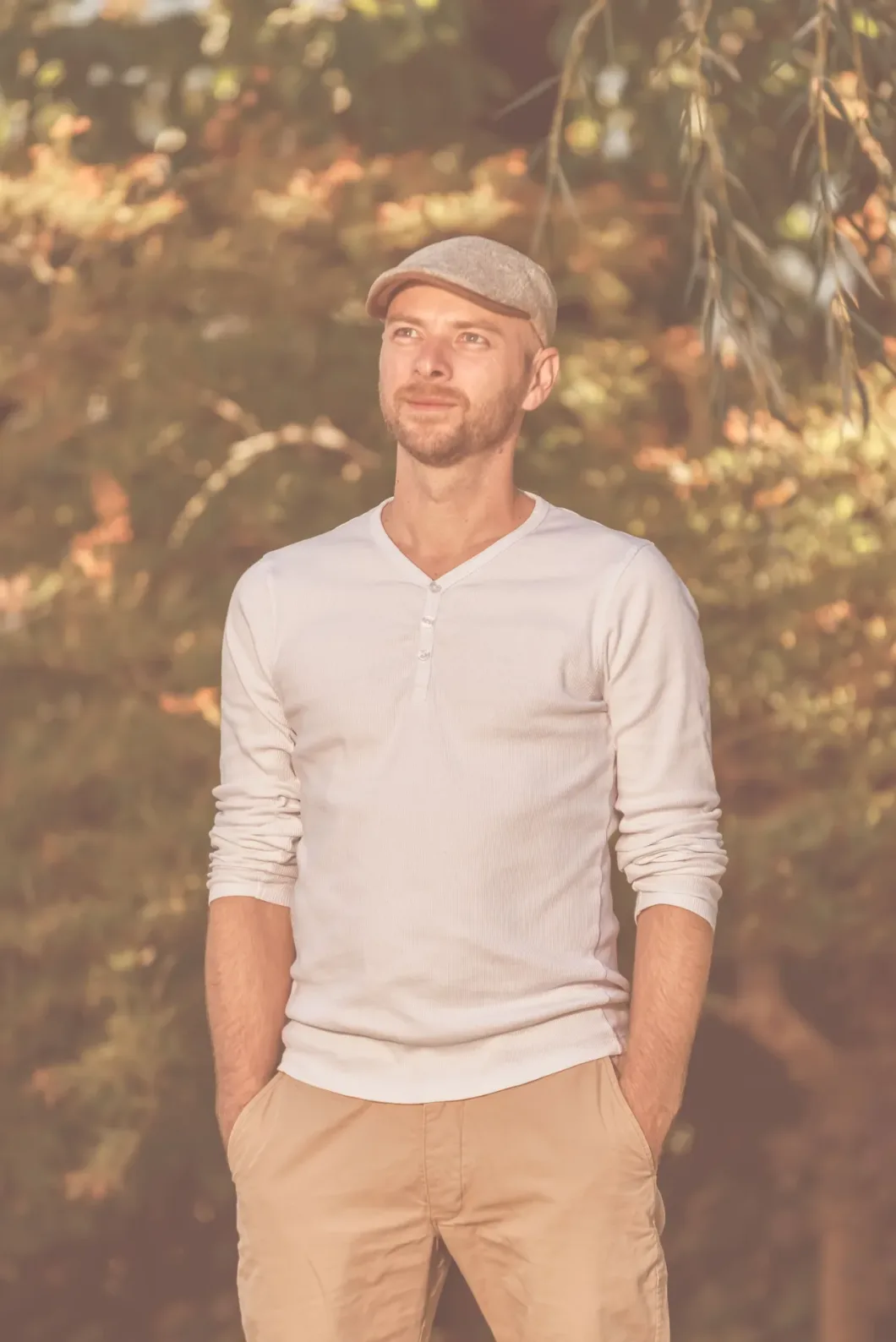 Mann mit Mütze im Freien, helles Langarmshirt, posiert entspannt im Grünen.
