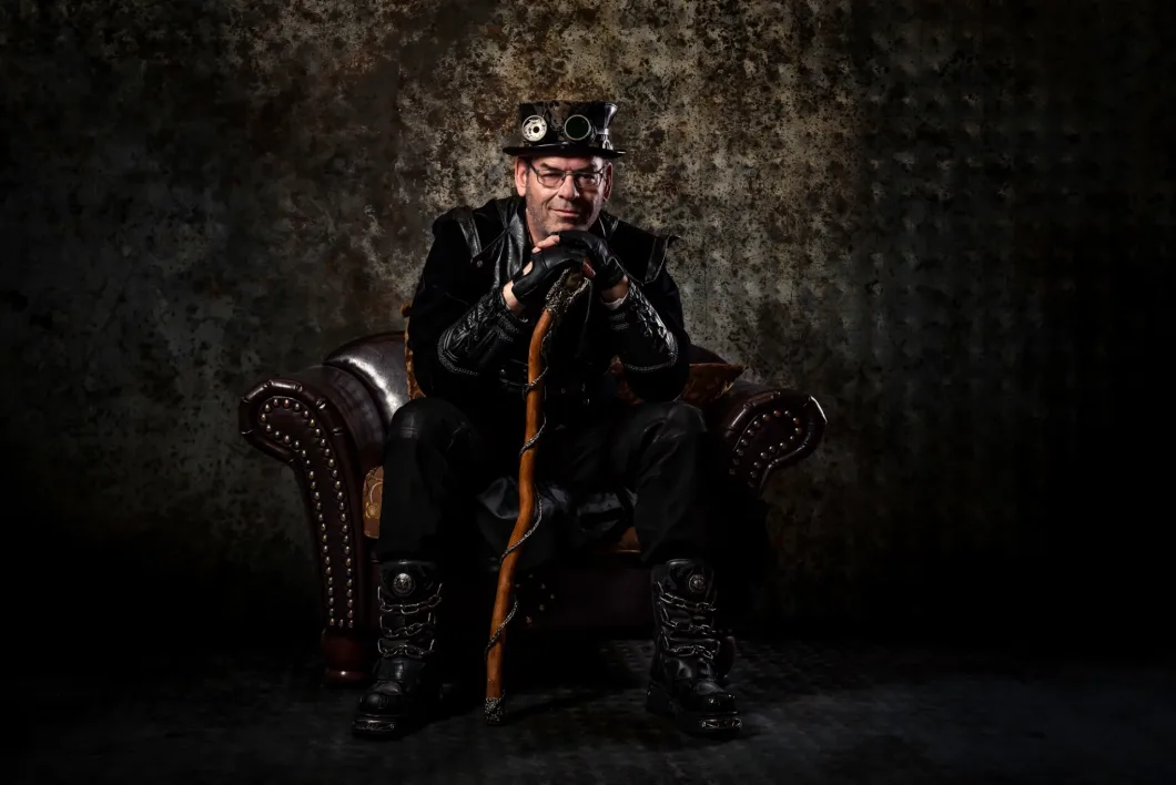 Mann in Steampunk-Kleidung sitzt auf Ledersessel, hält Gehstock, vor strukturiertem Hintergrund.