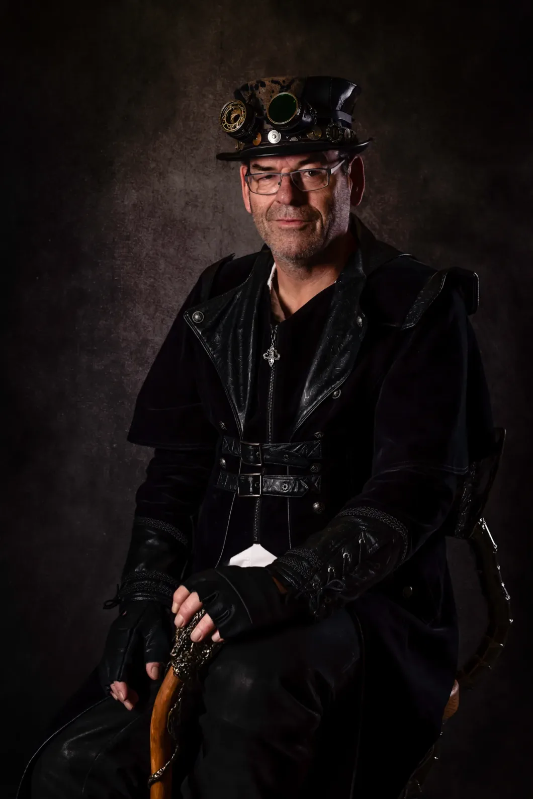Mann in Steampunk-Kleidung mit Hut und Handschuhen sitzt auf Hocker vor dunklem Hintergrund.