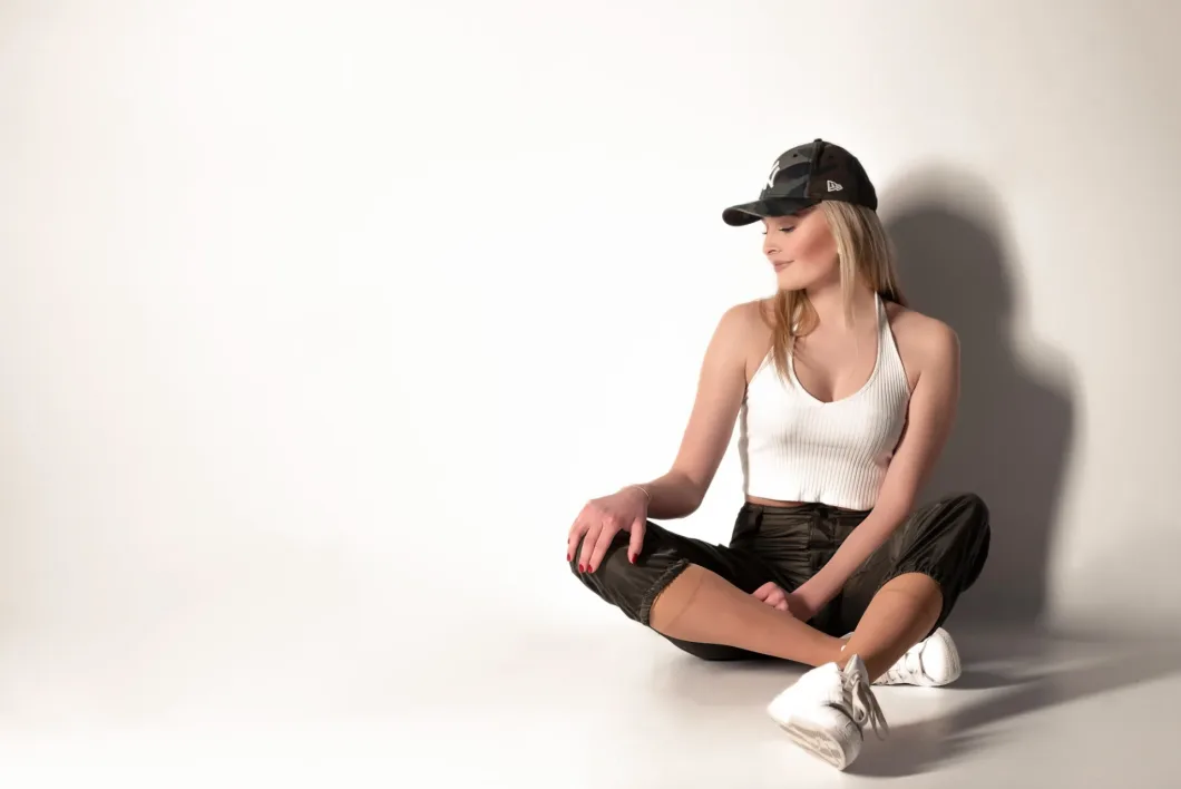 Frau sitzt lässig mit Cappy in weißem Top und schwarzen Hosen vor hellem Hintergrund.