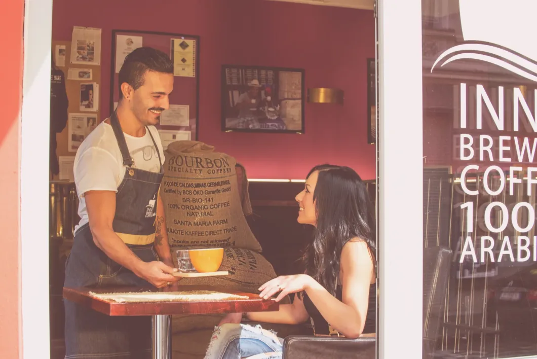 Barista serviert Kaffee einer sitzenden Frau in einem gemütlichen Café.