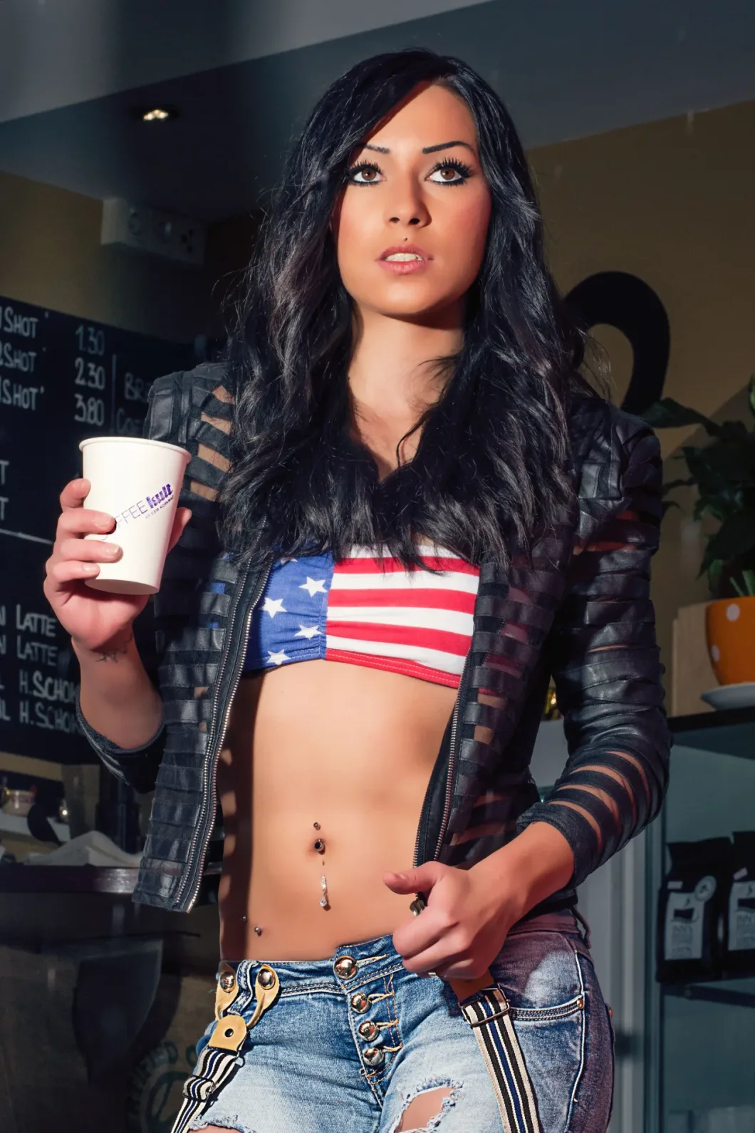 Frau in Lederjacke mit Kaffeebecher, trägt bauchfreies Top mit US-Flagge und Jeans.