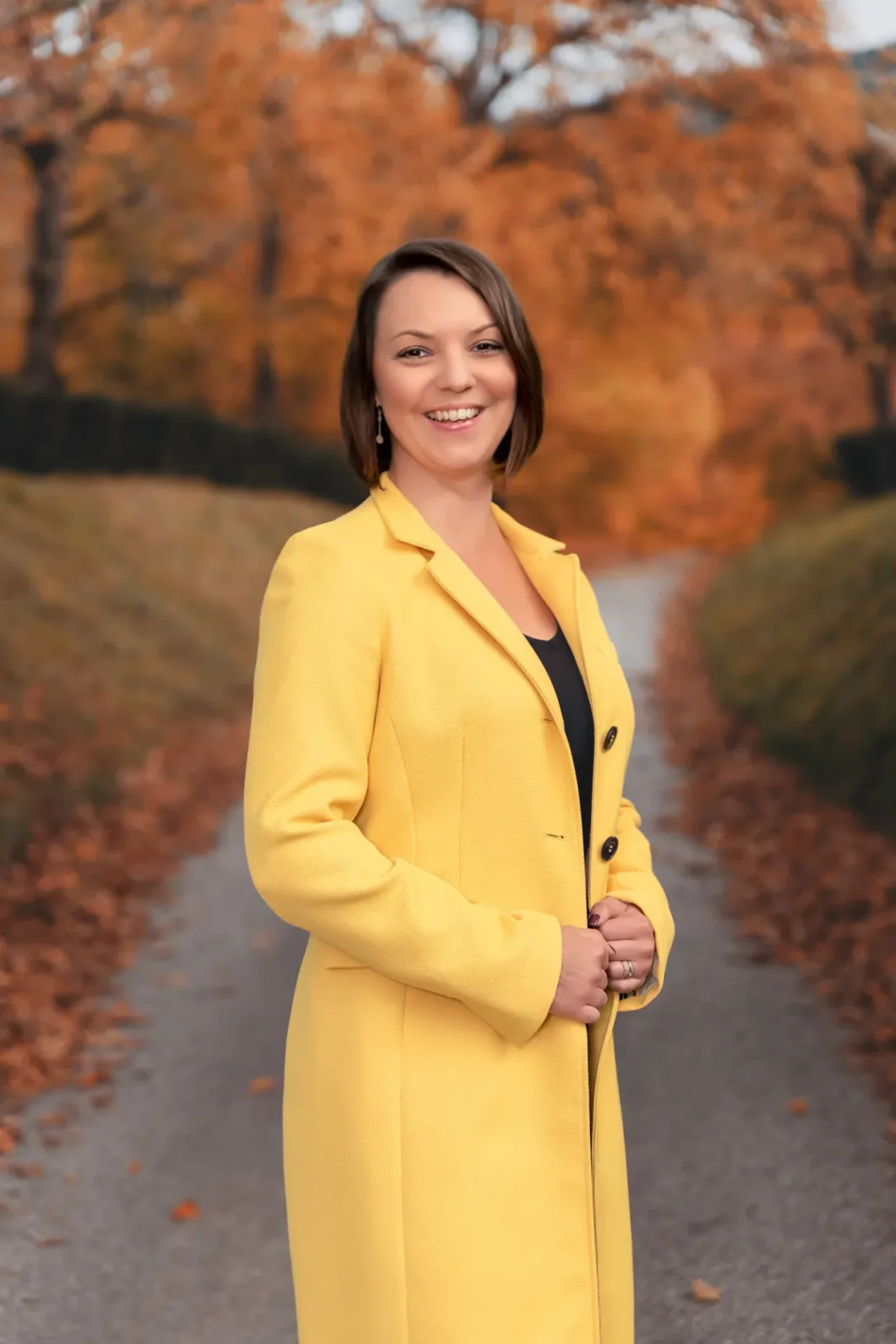 Frau in gelbem Mantel im herbstlichen Parkweg, lächelnd in die Kamera.