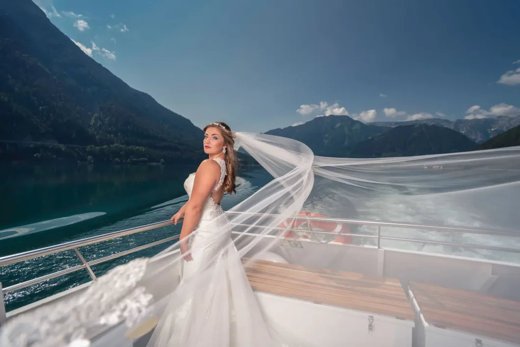 Braut in weißem Kleid mit Schleier auf einem Boot vor malerischer Bergkulisse.