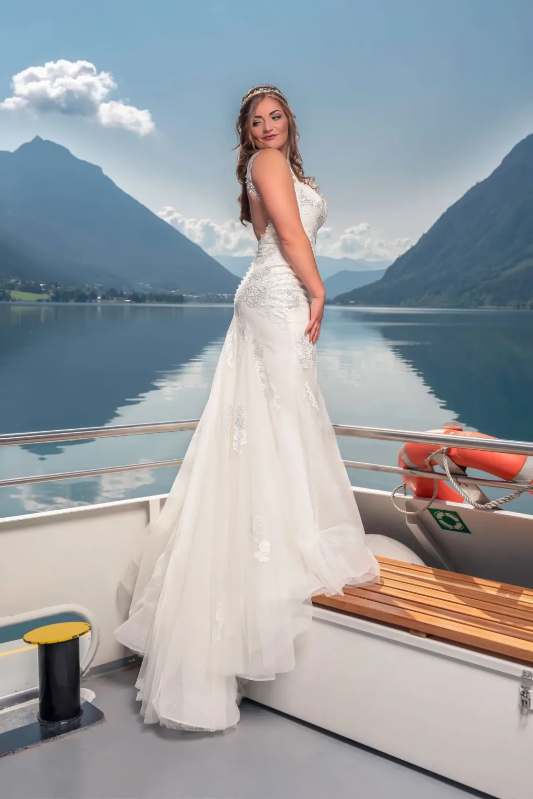 Braut in Spitze posiert auf Bootsdeck vor Bergkulisse und See unter blauem Himmel.