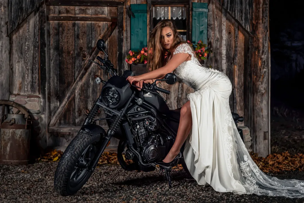 Frau im Hochzeitskleid posiert mit einem Motorrad vor einer rustikalen Holzwand.