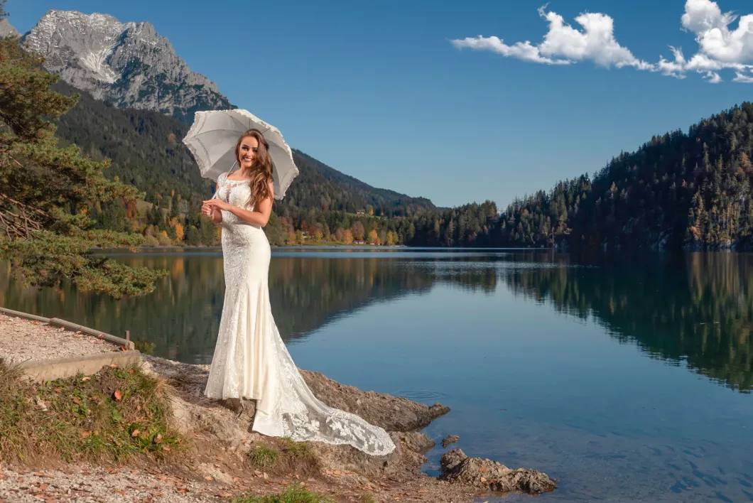 Braut in weißem Kleid mit Schirm vor Bergsee-Panorama mit blauem Himmel.