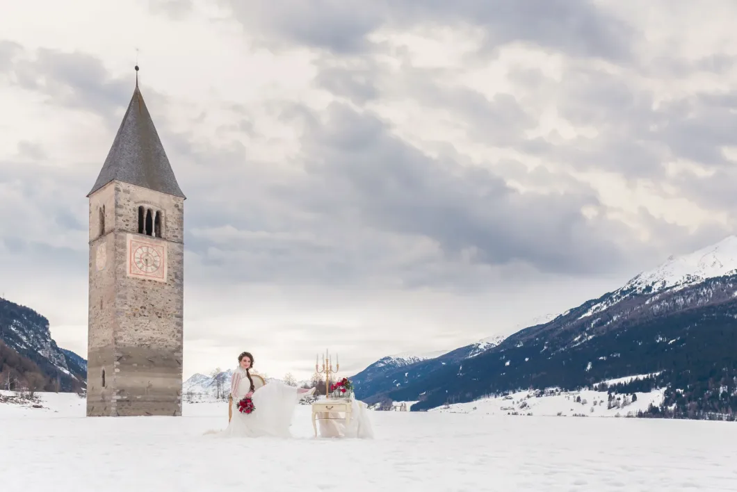 Frau in Brautkleid neben Turm im Schnee, Alpengipfel im Hintergrund.