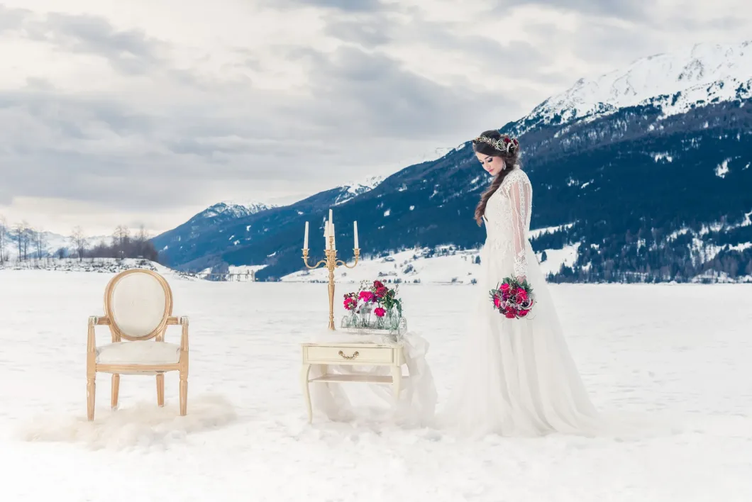 Braut im Schnee mit Blumen, neben Tisch und Kerzenhalter, Berglandschaft im Hintergrund.