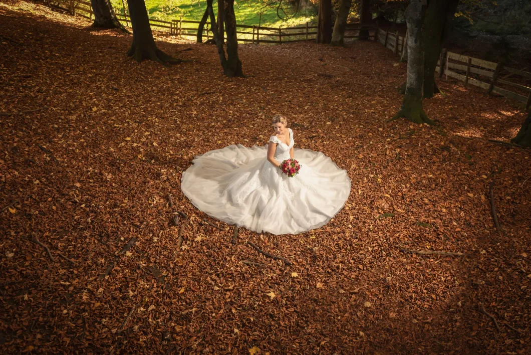 Braut sitzend im Laub mit weißem Kleid und Blumenstrauß, von oben fotografiert.