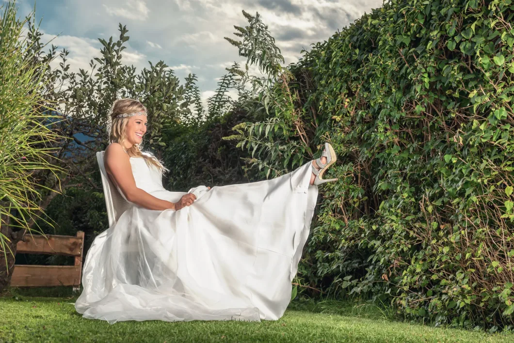 Braut in weißem Kleid sitzt lächelnd im Garten auf Holzstuhl, hebt ein Bein spielerisch an.