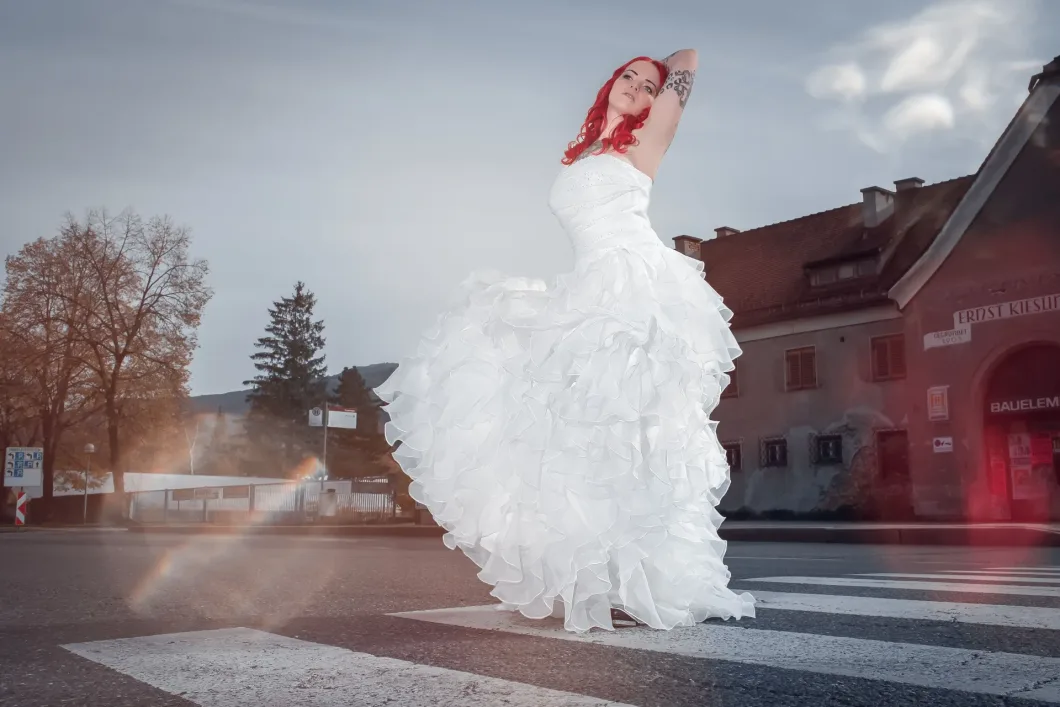 Frau in weißem Brautkleid mit roten Haaren auf Zebrastreifen, Gebäude im Hintergrund.