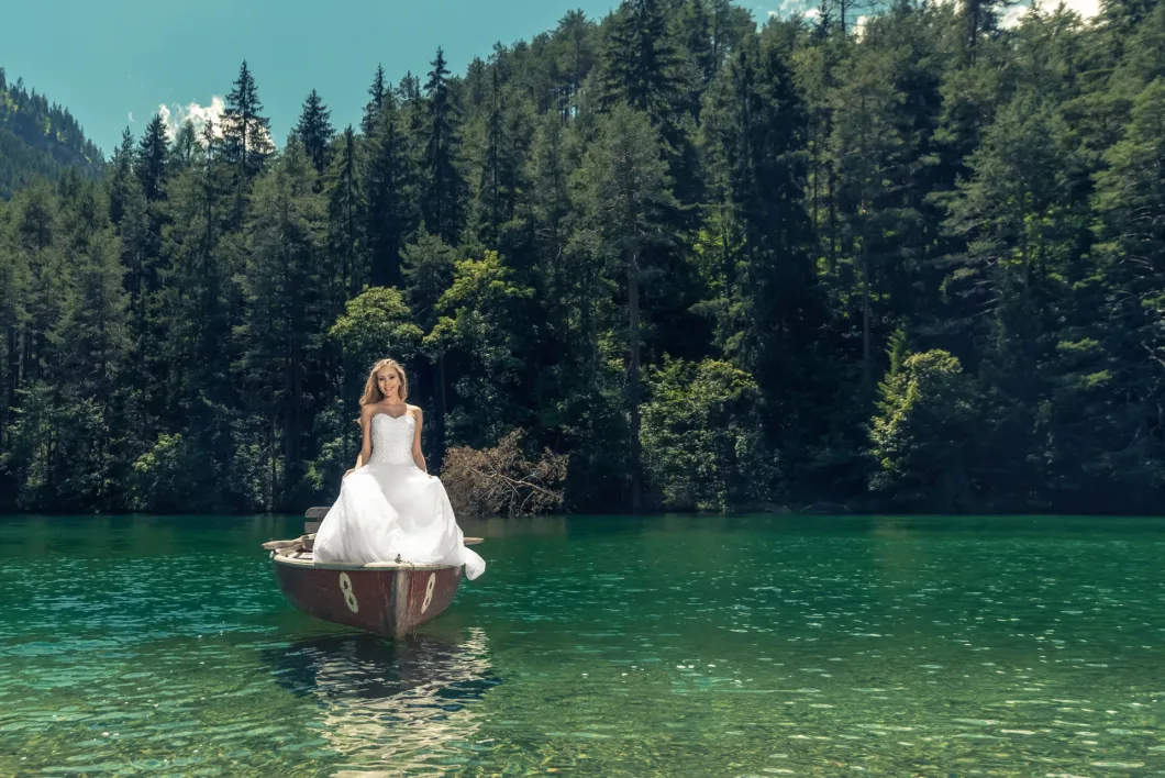 Braut im weißen Kleid auf einem Ruderboot, umgeben von Wald und klarem Wasser.