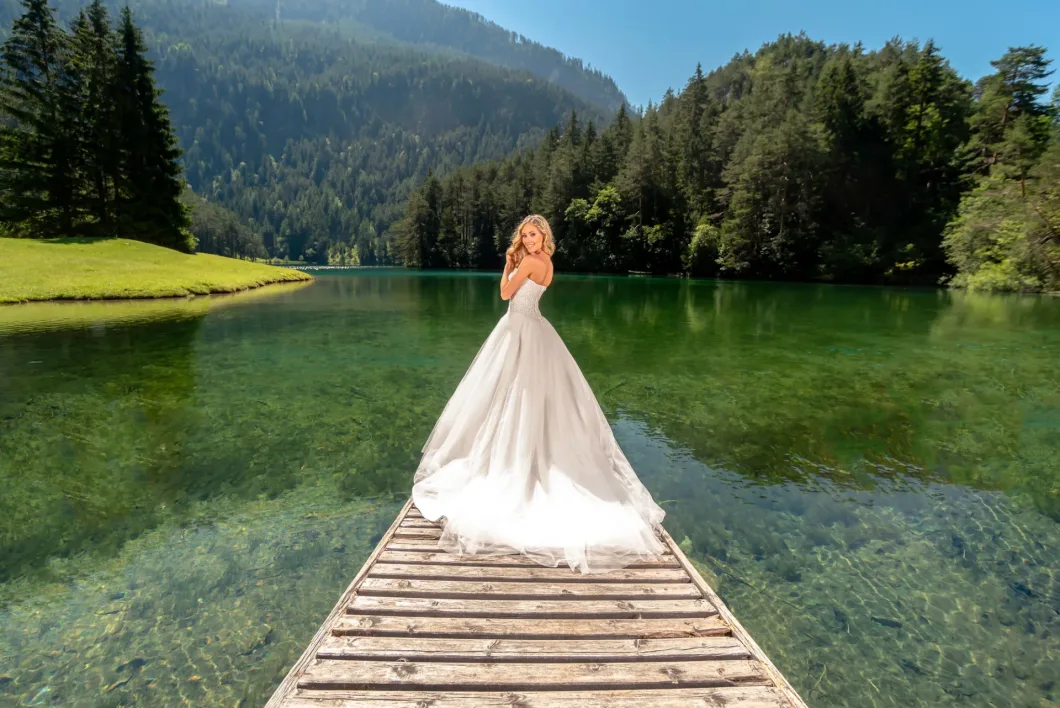 Braut im weißen Kleid auf Steg, umgeben von See und Wald. Klarer Himmel, sonniger Tag.