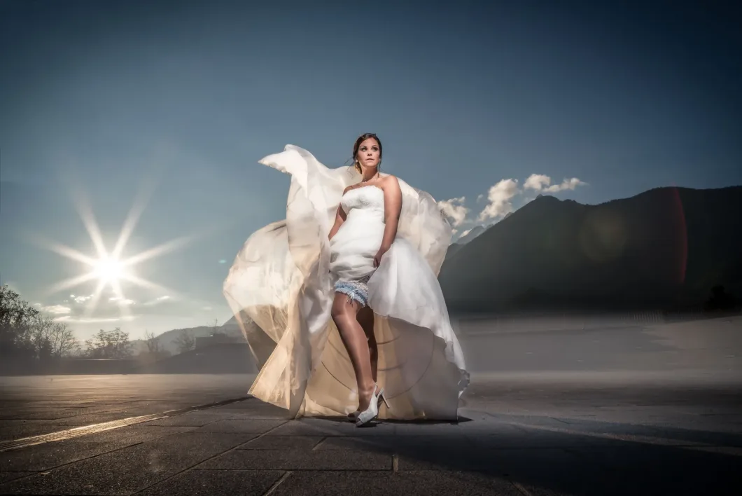 Braut in weißem Kleid mit fliegendem Stoff vor Berglandschaft bei Sonnenuntergang.