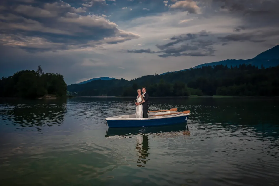 Brautpaar auf Ruderboot bei Sonnenuntergang auf See, umgeben von Bergen und bewölktem Himmel.