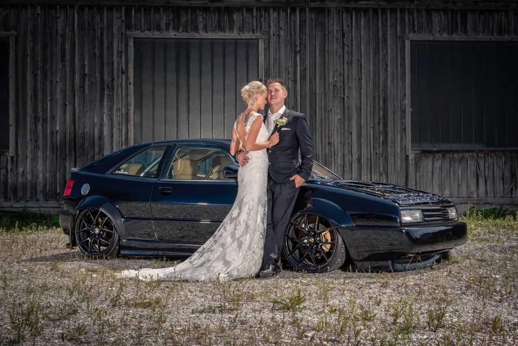 Paar in Hochzeitskleidung posiert vor elegantem schwarzen Auto auf Schotterplatz.