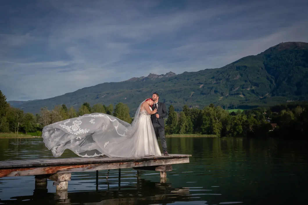 Brautpaar auf Bootssteg vor Berglandschaft, Brautkleid weht im Wind.