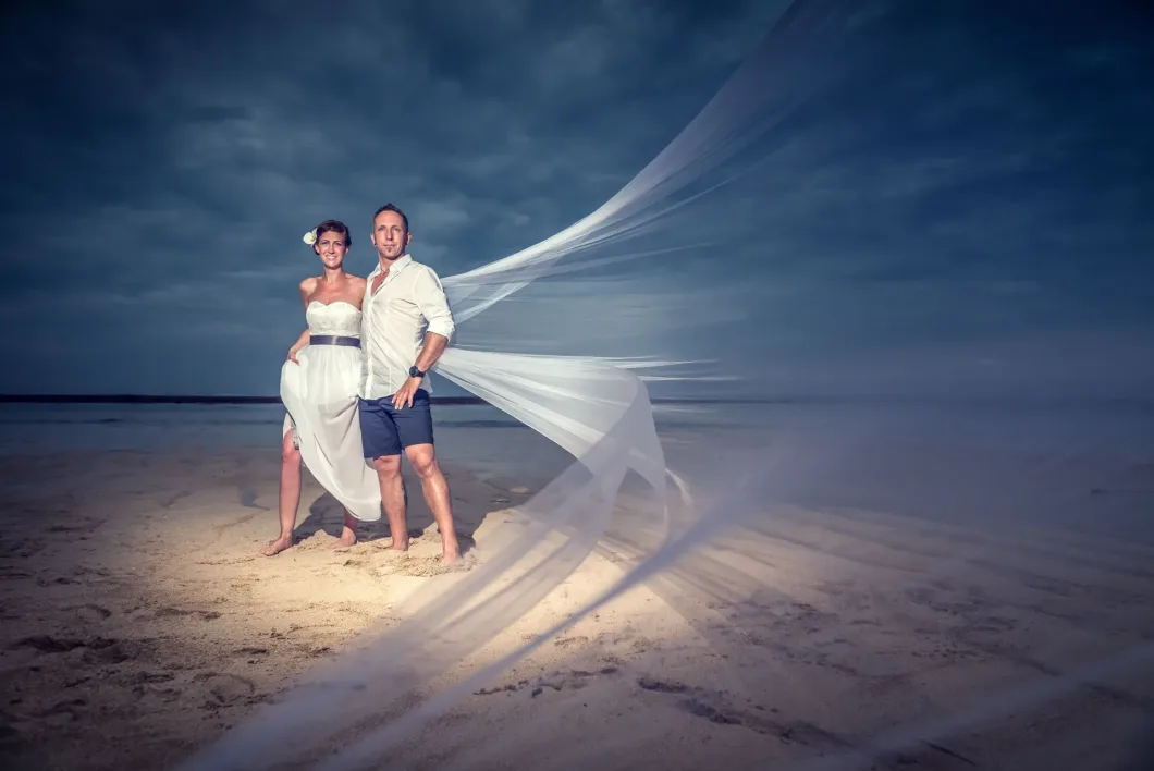 Brautpaar am Strand mit wehenden Schleiern vor dramatischem Himmel.