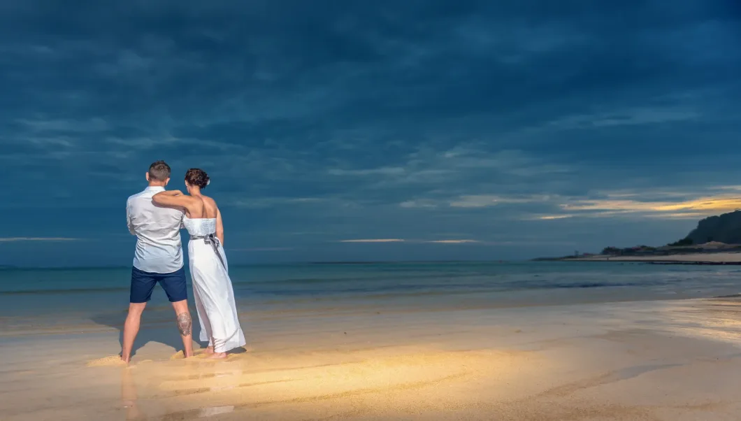 Paar am Strand bei Sonnenuntergang, Rücken zur Kamera, umarmt, romantische Stimmung.
