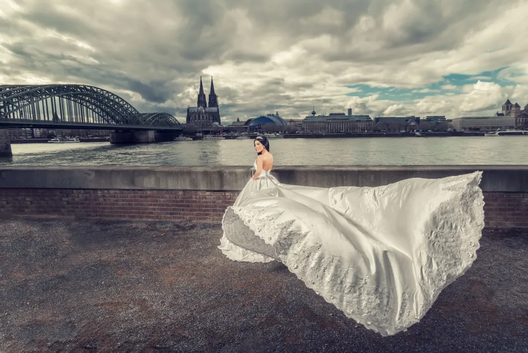 Braut in elegantem weißen Kleid vor Rhein und Kölnpanorama mit Wolkenhimmel.