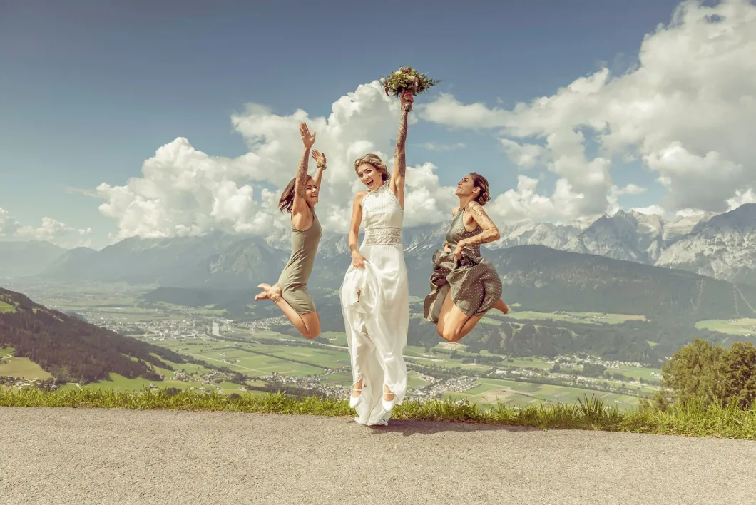 Braut und zwei Frauen springen freudig vor Alpenpanorama in die Luft.