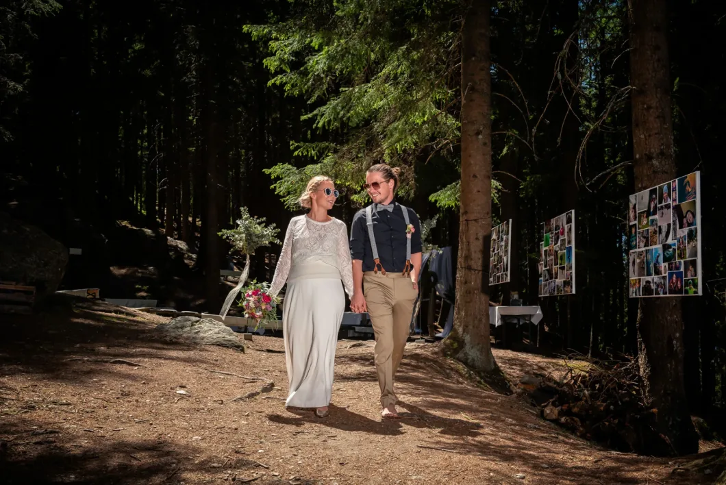 Brautpaar spaziert lächelnd im Wald. Hochzeitsfotos sind im Hintergrund sichtbar.