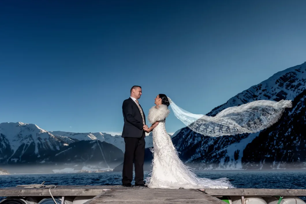 Brautpaar auf Steg vor Bergkulisse, Hochzeitskleid mit Schleier, klarer Himmel, winterliche Landscha