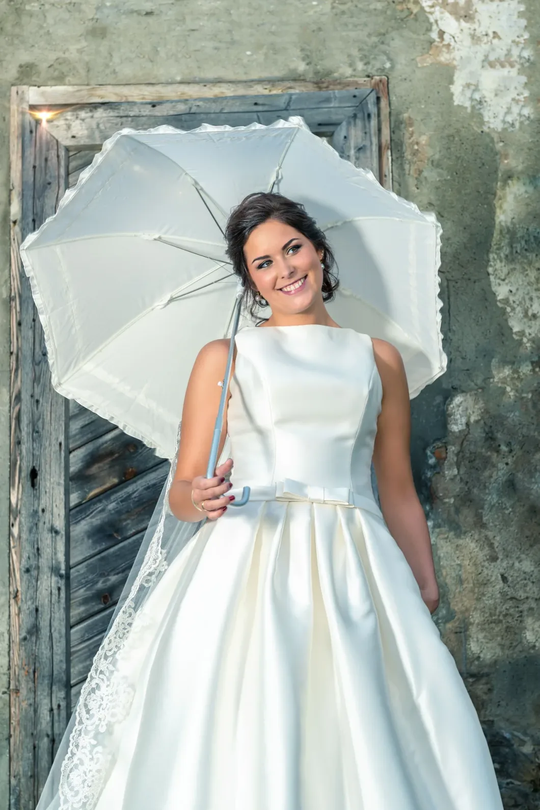 Braut in weißen Kleid mit Schirm vor einer alten Holztür mit verwitterter Mauer.