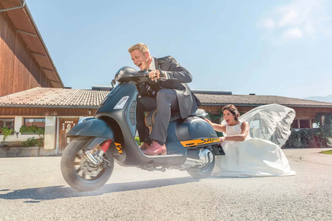 Bräutigam fährt Vespa, Braut hält sich am schleifenden Kleid fest.
