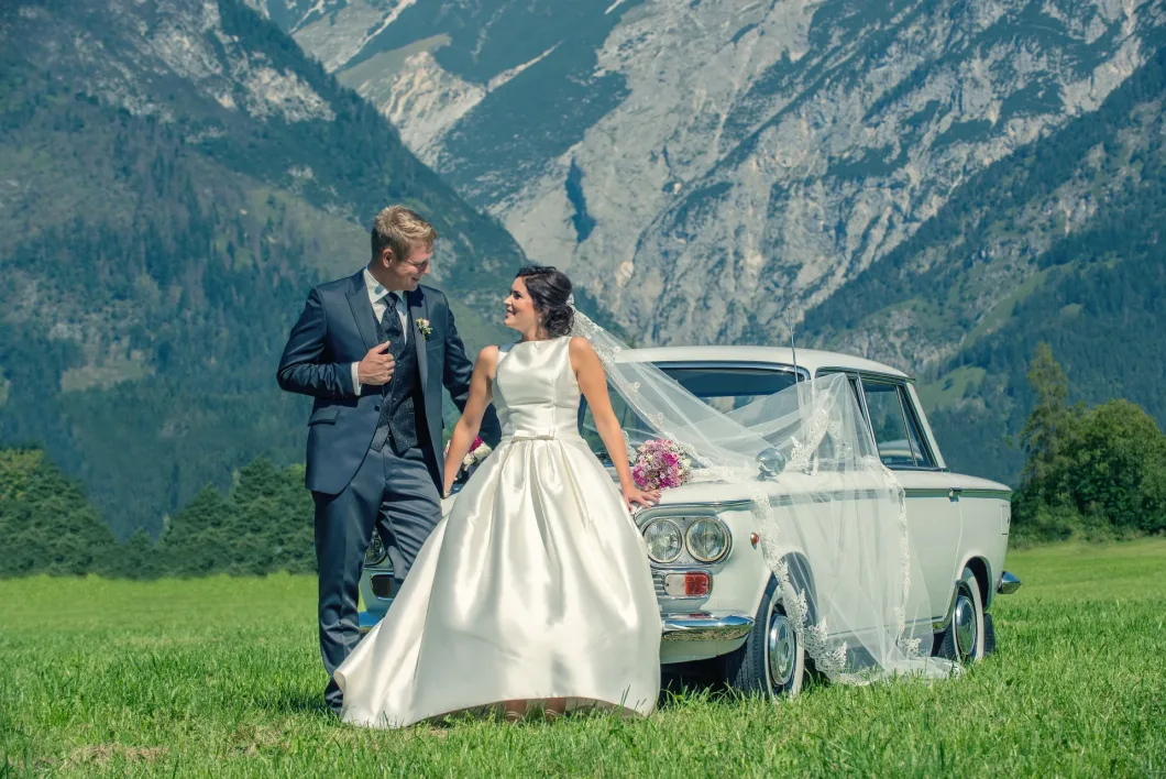 Brautpaar vor klassischem Auto in Berglandschaft steht lachend auf einer Wiese.