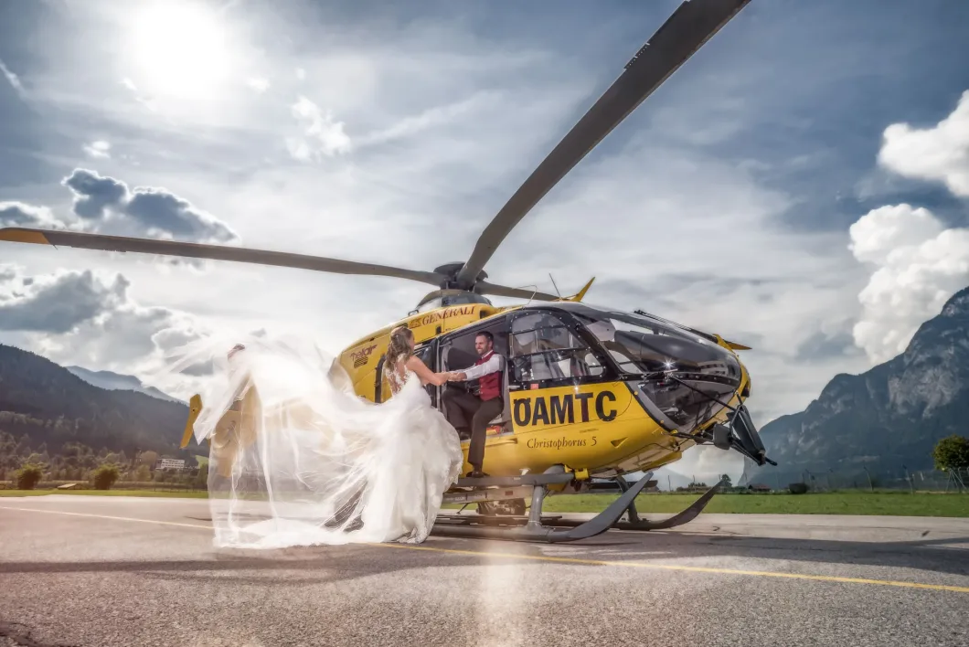 Brautpaar vor Rettungshubschrauber, Brautschleier weht im Wind, Alpenlandschaft im Hintergrund.