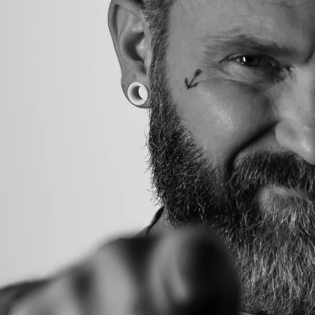 Mann mit Bart und Ohrschmuck zeigt auf Kamera, Pfeiltattoo unter Auge. Schwarz-Weiß.